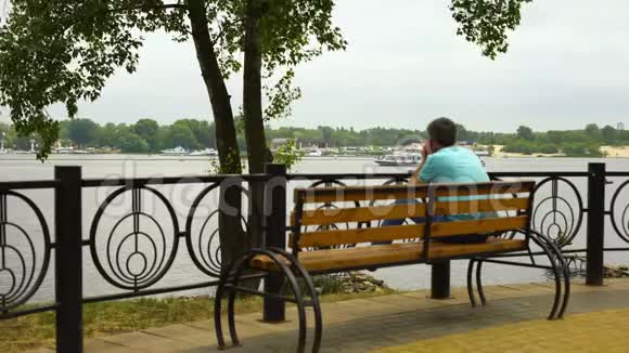 男子坐在城市长椅上欣赏公园的河景游船经过视频的预览图