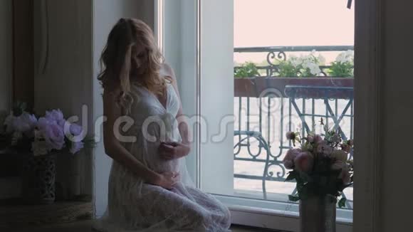 穿着白色连衣裙的孕妇正坐在窗边穿着白色蕾丝连衣裙从上面看幸福的女人期待着视频的预览图