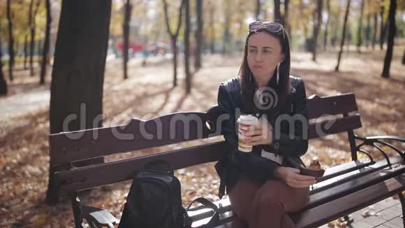 在秋季公园的长椅上吃午饭迷人的女孩从纸杯里喝咖啡吃羊角面包视频的预览图