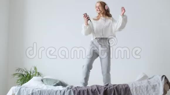 戴着耳机听音乐和在室内床上跳跃的年轻休闲女子视频的预览图