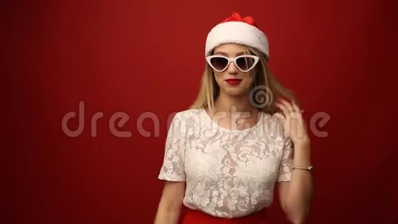 戴着圣诞老人帽子戴着墨镜的迷人金发女郎红色的背景特写肖像视频的预览图