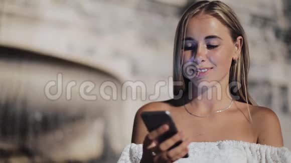 迷人的年轻女性使用智能手机坐在夜城喷泉附近的街道上对这条信息做出了愉快的反应视频的预览图