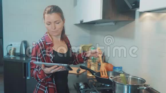 女人妈妈在厨房做饭的概念在平底锅里煮汤看看数字平板电脑上的食谱女孩视频的预览图