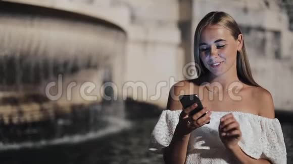 迷人的年轻女性使用智能手机坐在夜城喷泉附近的街道上对这条信息做出了愉快的反应视频的预览图