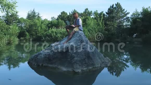 一个年轻的自由职业者坐在湖中央的石头上手提电脑自由职业概念出差工作无线无线视频的预览图