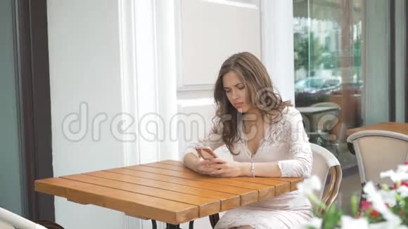 悲伤的年轻美丽的女孩坐在街头咖啡馆在电话上写短信视频的预览图