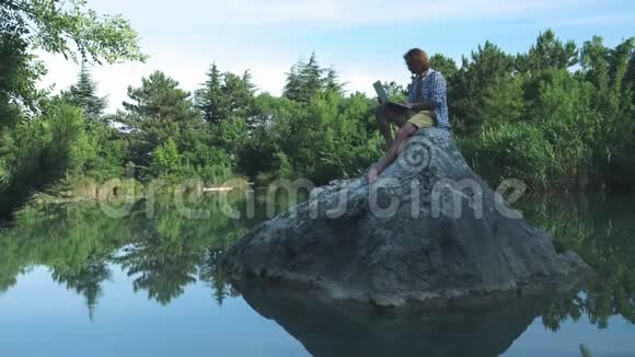 一个年轻的自由职业者坐在湖中央的石头上手提电脑自由职业概念出差工作无线无线视频的预览图