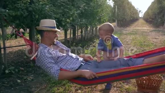 在苹果花园笑的孩子在水果篮附近的吊床上摇着他的父亲慢动作视频的预览图