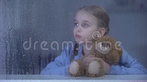 小女孩抱着泰迪熊在雨天透过窗户看着视频的预览图