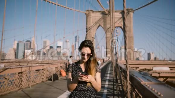 美丽的高加索女商人戴着时尚的太阳镜站在布鲁克林大桥4K使用智能手机信使应用程序视频的预览图