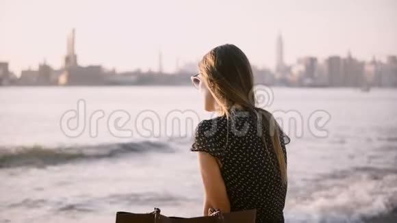 美丽的白种人女孩戴着太阳镜独自坐在城市海滩上在令人惊叹的纽约日落4K上回顾相机视频的预览图