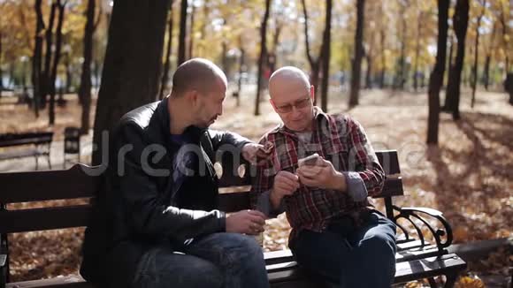 一位年迈父亲的成年儿子在秋公园的长凳上教智能手机视频的预览图