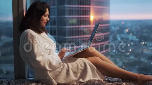 穿着浴袍的年轻女子坐在酒店高层的窗户附近在笔记本电脑前休息的女孩视频的预览图