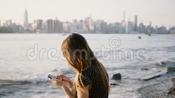 无法辨认的女人长发使用智能手机应用程序在河滩与纽约城市天际线景观日落4K视频的预览图
