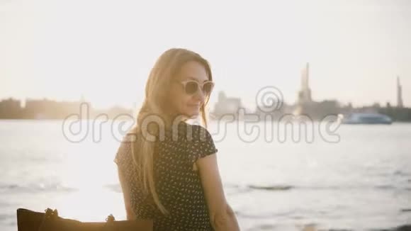 美丽的欧洲女人戴着太阳镜回头看镜头转身离开阳光明媚的城市海滩享受夏日日落4K视频的预览图