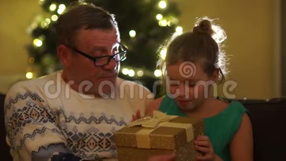 祖父和孙女靠近圣诞树女孩打开礼物高兴起来新年礼物视频的预览图