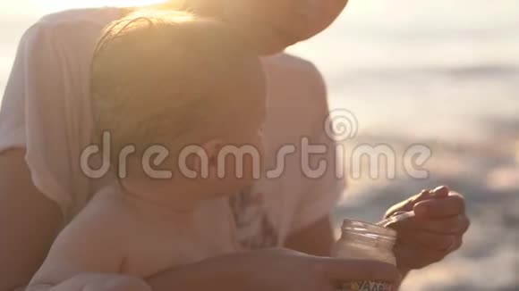妈妈在日落时用勺子从靠近海洋的海滩上的罐子里喂孩子视频的预览图