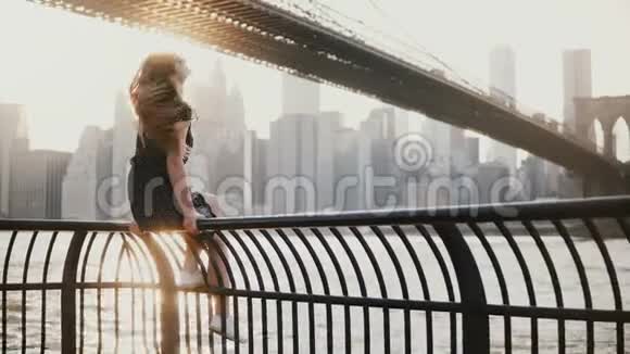 美丽的年轻女孩戴着太阳镜坐在纽约布鲁克林大桥附近的河堤围栏上欣赏4K风景视频的预览图