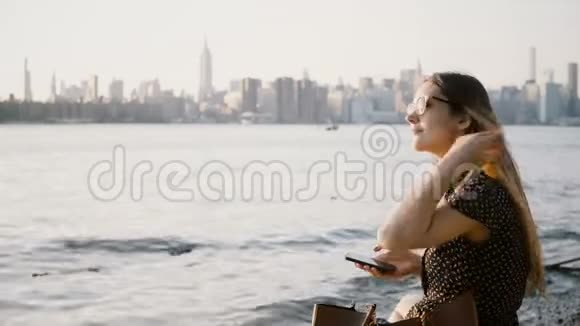 美丽的欧洲女孩长头发戴太阳镜使用智能手机购物在线应用程序在纽约日落海滩4K视频的预览图