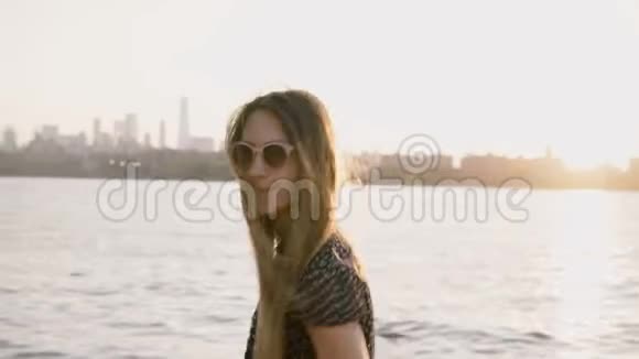 快乐的年轻欧洲女孩的肖像戴着太阳镜摆姿势头发随风飘动令人惊叹的纽约日落4K视频的预览图