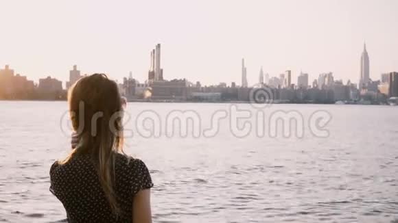 戴着太阳镜的漂亮女孩独自站着头发随风飘扬在纽约日落4K时走到河边视频的预览图