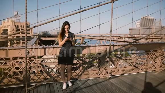 快乐的欧洲年轻女子使用智能手机然后打电话和朋友在布鲁克林大桥纽约4K视频的预览图