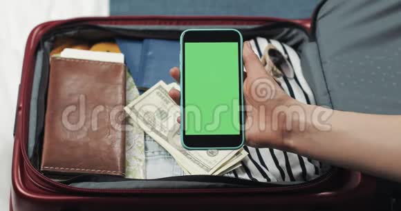 年轻女子正在准备旅行她拿着智能手机手提箱背景上有绿色屏幕铬钥匙视频的预览图