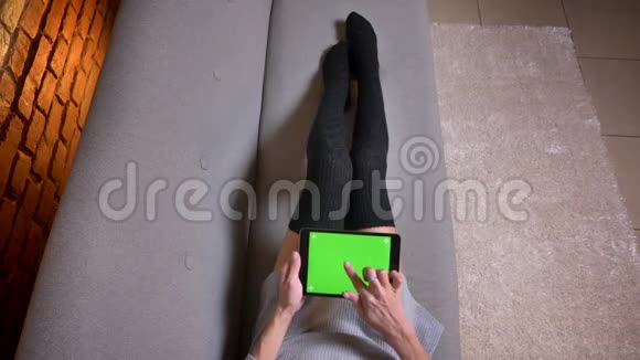室内沙发上穿着可爱袜子的女腿特写镜头女人用绿色屏幕浏览平板电脑视频的预览图