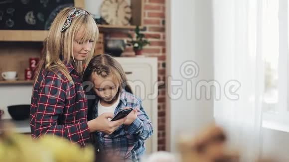 快乐的母亲和女儿一起浏览智能手机早上在家厨房微笑视频的预览图