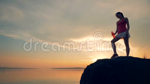 女跑步者喝水慢动作一个女人站在靠近水的岩石上在夕阳的背景下喝酒视频的预览图