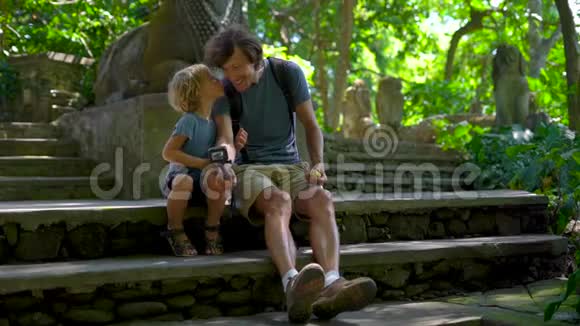 一个年轻人和他的小儿子坐在Ubud的猴子森林生态公园的楼梯上慢镜头视频的预览图