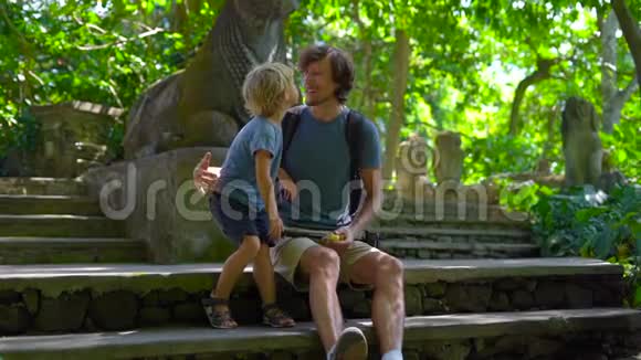 一个年轻人和他的小儿子坐在Ubud的猴子森林生态公园的楼梯上慢镜头视频的预览图