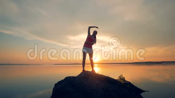 女运动员在湖边的岩石上热身一个女孩在夕阳背景下奔跑之前伸展身体视频的预览图