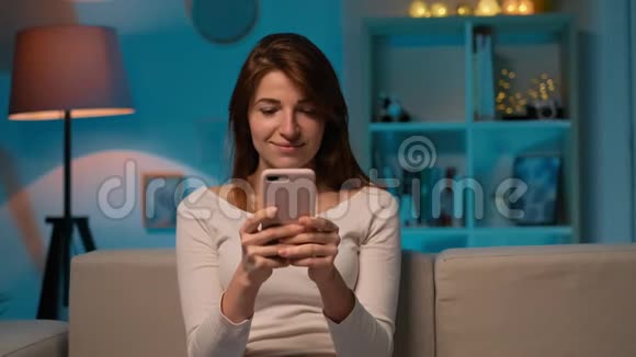 年轻女子坐在沙发上用手机聊天在一个舒适的房间里大笑室内女孩视频的预览图