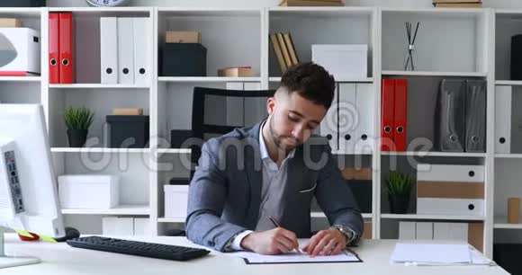 穿着灰色夹克的商人坐在白色办公室的桌子旁签名敲敲和扔一张纸视频的预览图
