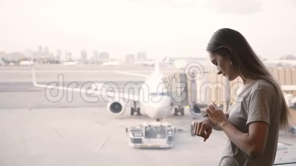 年轻的高加索女商人站在机场候机室的窗口用智能手表看着飞机视频的预览图