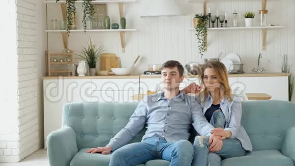 一对夫妇坐在舒适的蓝色沙发上在现代厨房里看电视视频的预览图