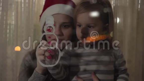 妈妈和女儿在窗玻璃上画一个画着颜料的雪人用丙烯酸在玻璃上画画视频的预览图
