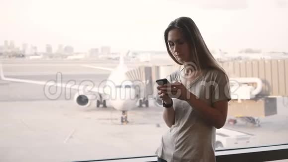 站在机场候机楼窗口附近的年轻欧洲妇女看着电话和智能手表感到沮丧和沮丧视频的预览图