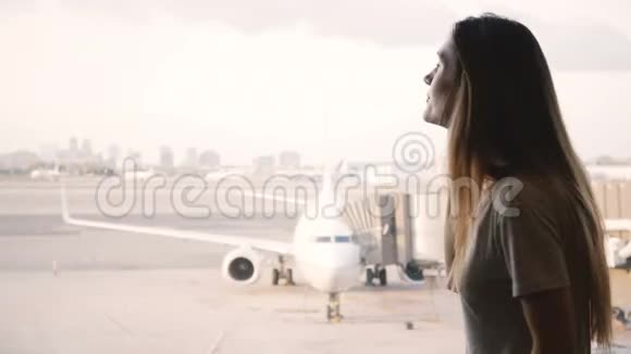 年轻的欧洲女孩在智能手机上交谈微笑着结束在机场航站楼窗口打电话飞机在后台视频的预览图