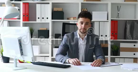 年轻男子灰色夹克坐在白色办公室的桌子上提供文件视频的预览图
