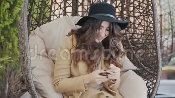 一位身穿外套头戴黑帽子的年轻美女坐在咖啡馆的椅子上在咖啡馆里打短信视频的预览图