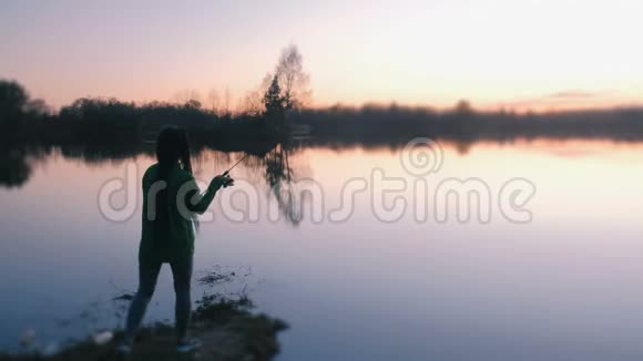 傍晚日落时分一个面目全非的女人在池塘的岸边钓鱼后景视频的预览图