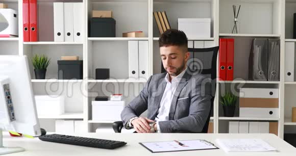 穿着灰色夹克的年轻商人坐在白色办公室的桌子上看着时钟坐在椅子上视频的预览图