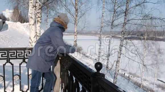 穿着蓝色羽绒服带着毛皮帽的人站在栅栏旁在冬季公园里环顾四周视频的预览图