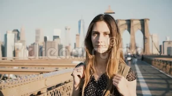 年轻的积极的女性白种人游客带着背包看着相机微笑着在阳光明媚的布鲁克林大桥纽约4K视频的预览图
