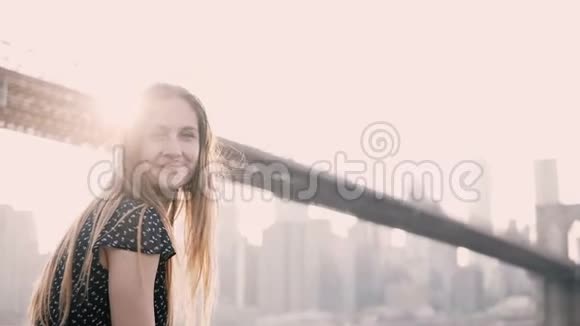 迷人的白种人女孩看着镜头微笑坐在堤岸围栏纽约布鲁克林大桥日落景色视频的预览图