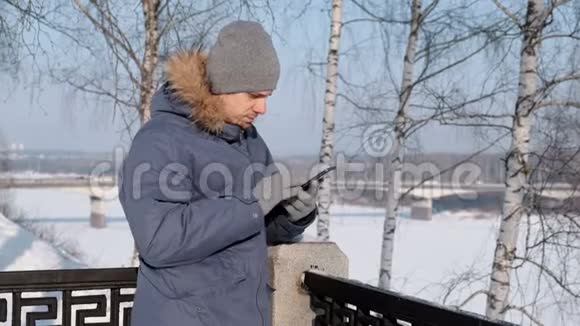 穿着蓝色羽绒服带着皮帽的人用手机上网在冬季公园聊天视频的预览图