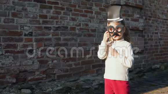 小女孩拿着纸胡子和帽子在砖墙旁边的棍子上视频的预览图