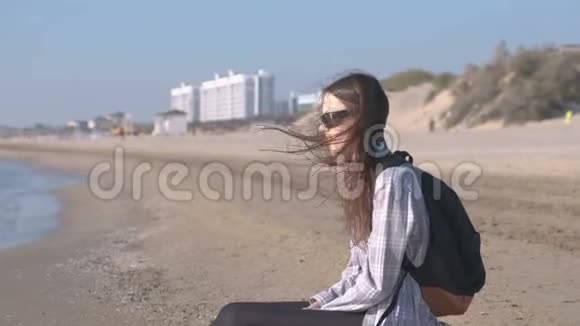 旅行者带着背包坐在沙滩上看着大海海滩上的年轻女孩视频的预览图